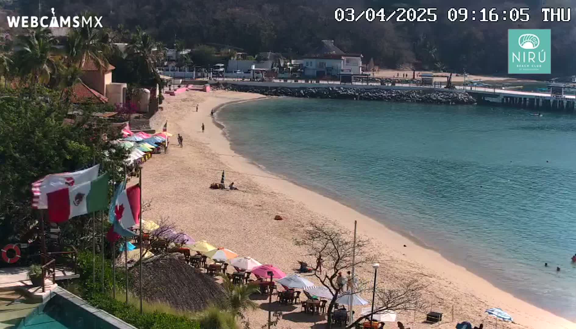 A sandy beach with calm, clear turquoise water, umbrellas, and a few people is seen on a sunny day, next to buildings and a small pier.