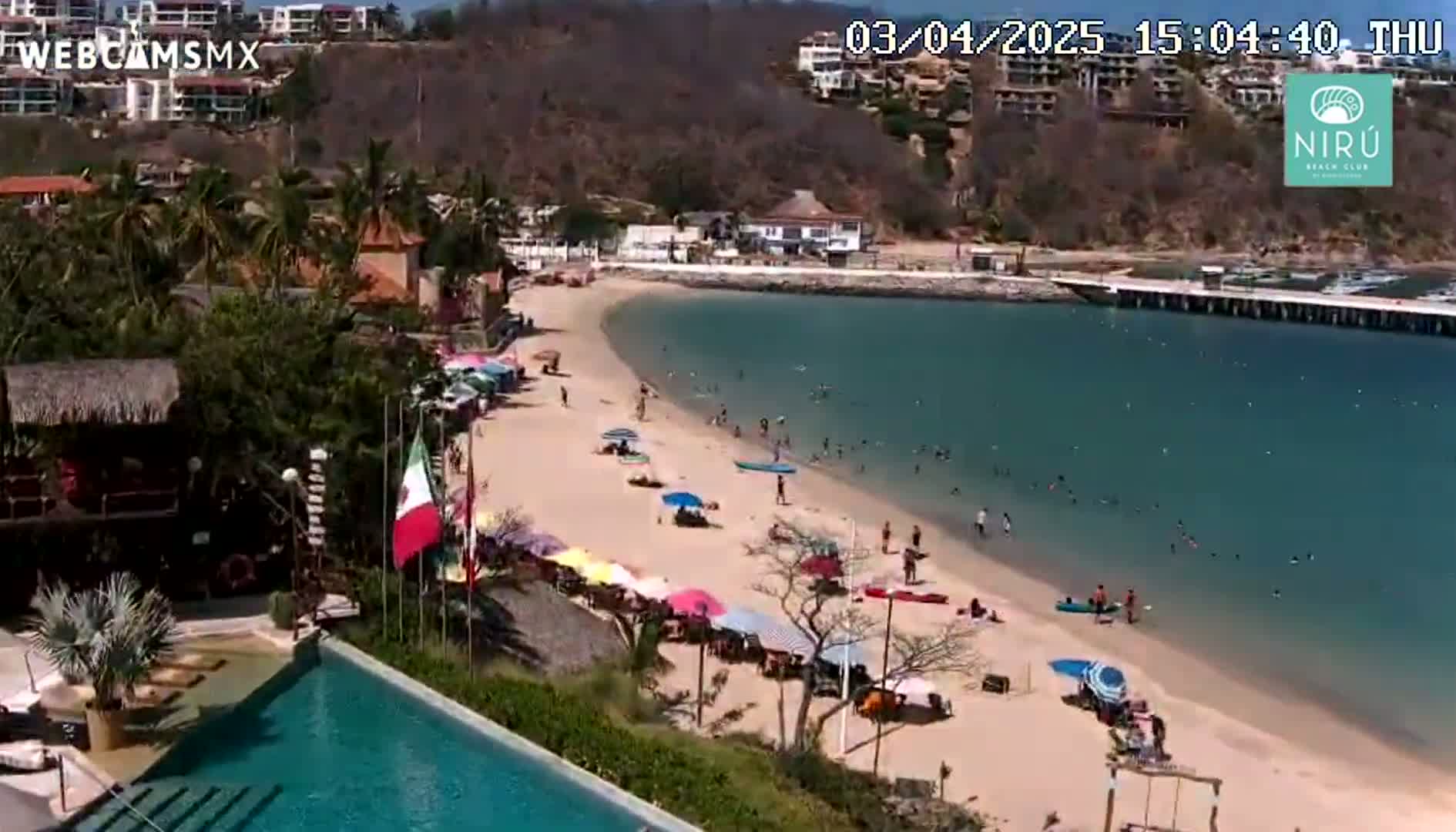 A sandy beach with numerous colorful umbrellas and people swimming in clear, calm water, overlooks a swimming pool and buildings nestled on a hillside under a sunny sky.