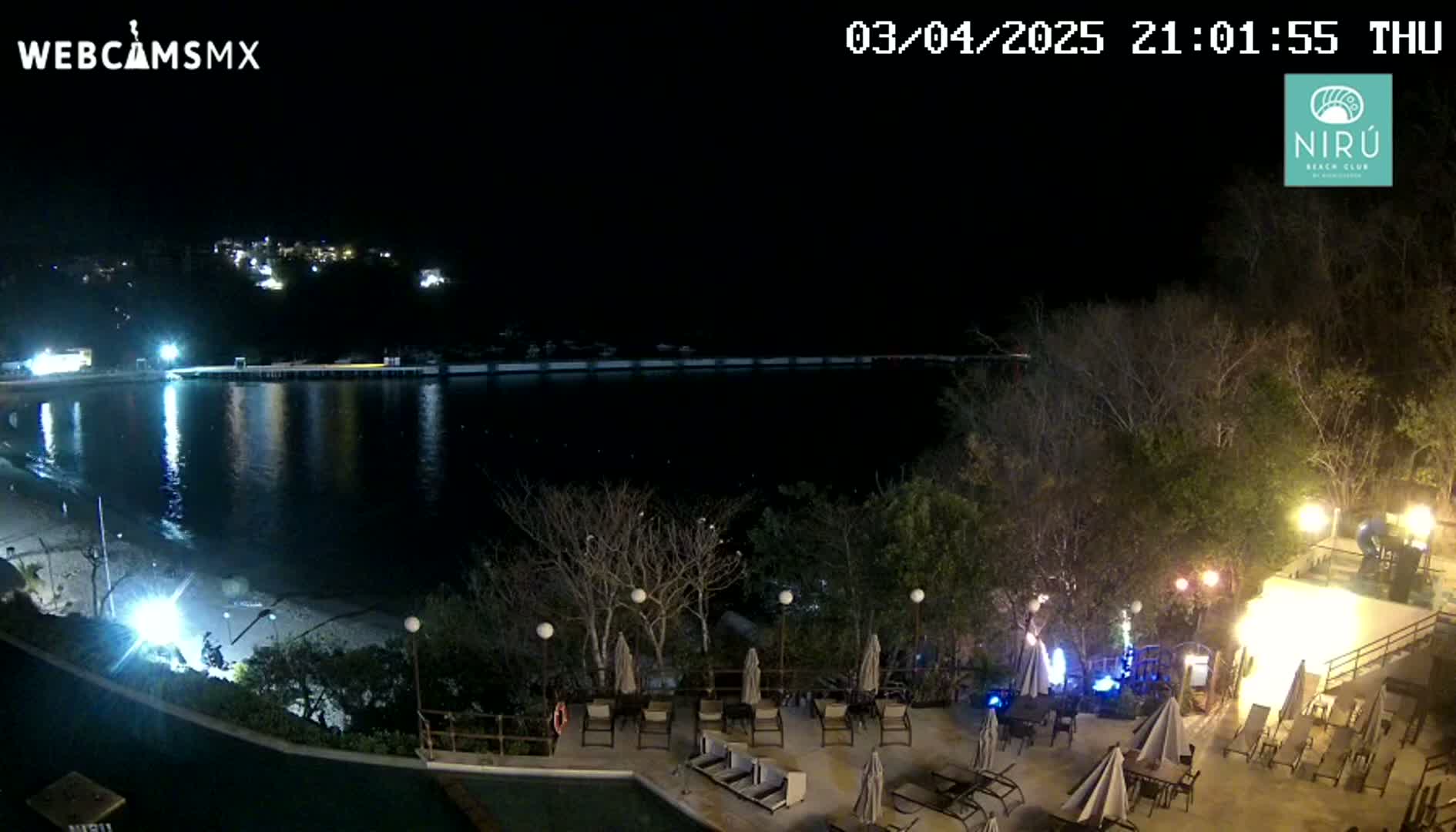 A nighttime view of a calm body of water next to a beach with outdoor furniture and lights at a resort.