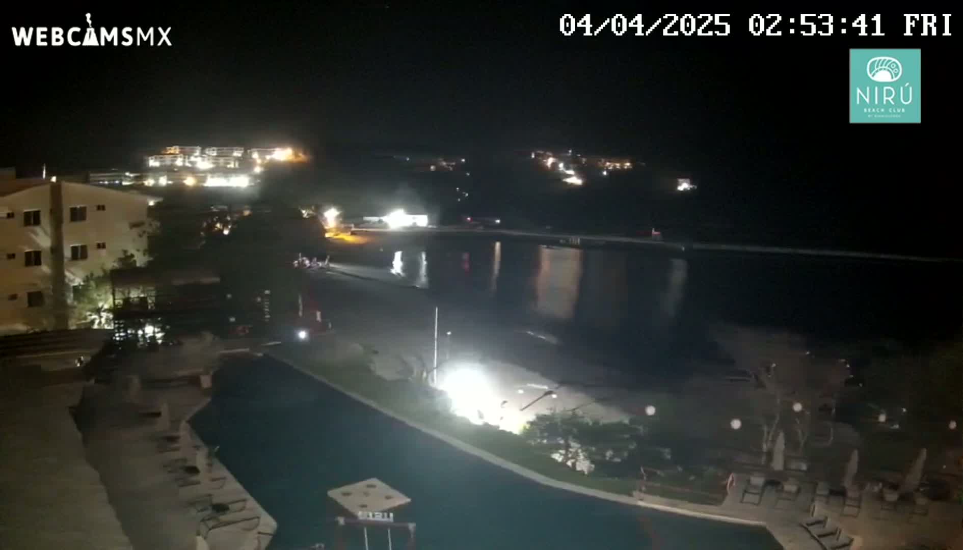 A nighttime view of a resort area near a body of water, showing buildings, a pool, and calm water reflecting lights.