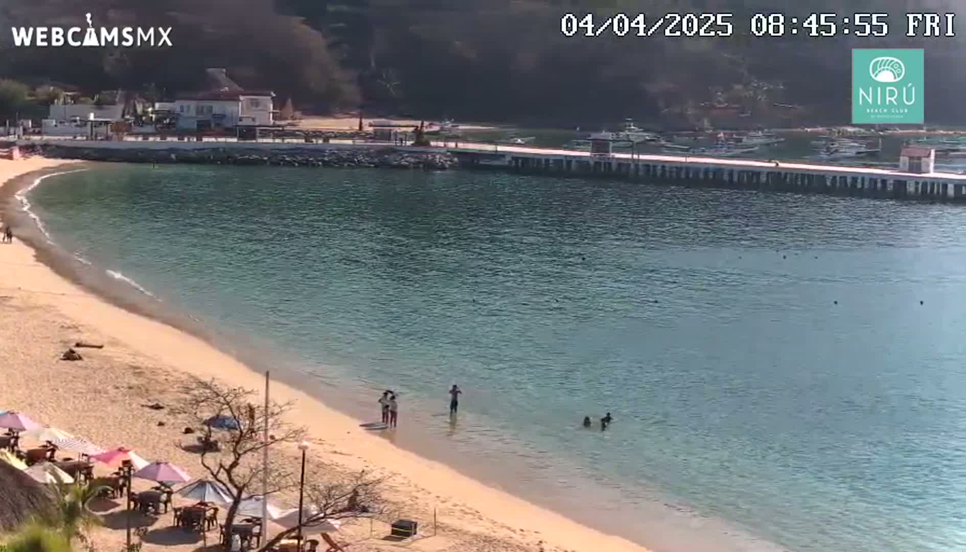 A sandy beach with calm, clear water laps the shore near a pier with several boats, and a few people are swimming and relaxing on the beach under a sunny sky.