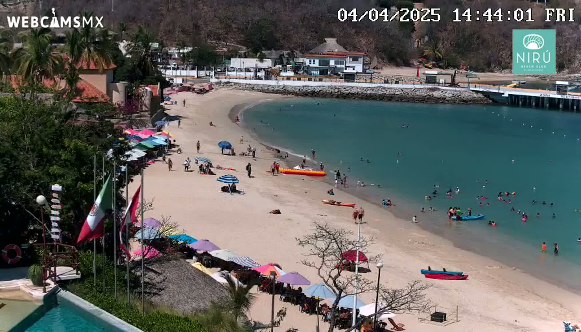 A sandy beach with numerous colorful umbrellas and people swimming and relaxing in calm, clear turquoise water, under a sunny sky.