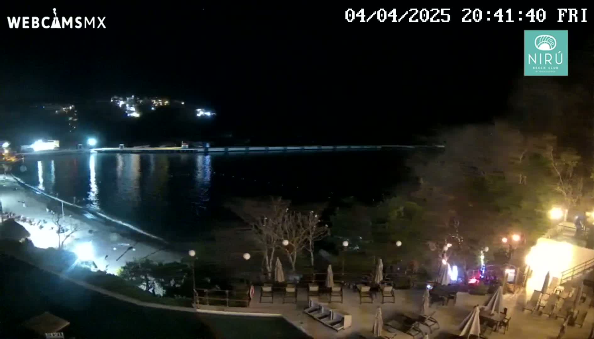A nighttime view of a calm, dark body of water reflecting lights from a shoreline with a beach, some buildings, and outdoor patio furniture.