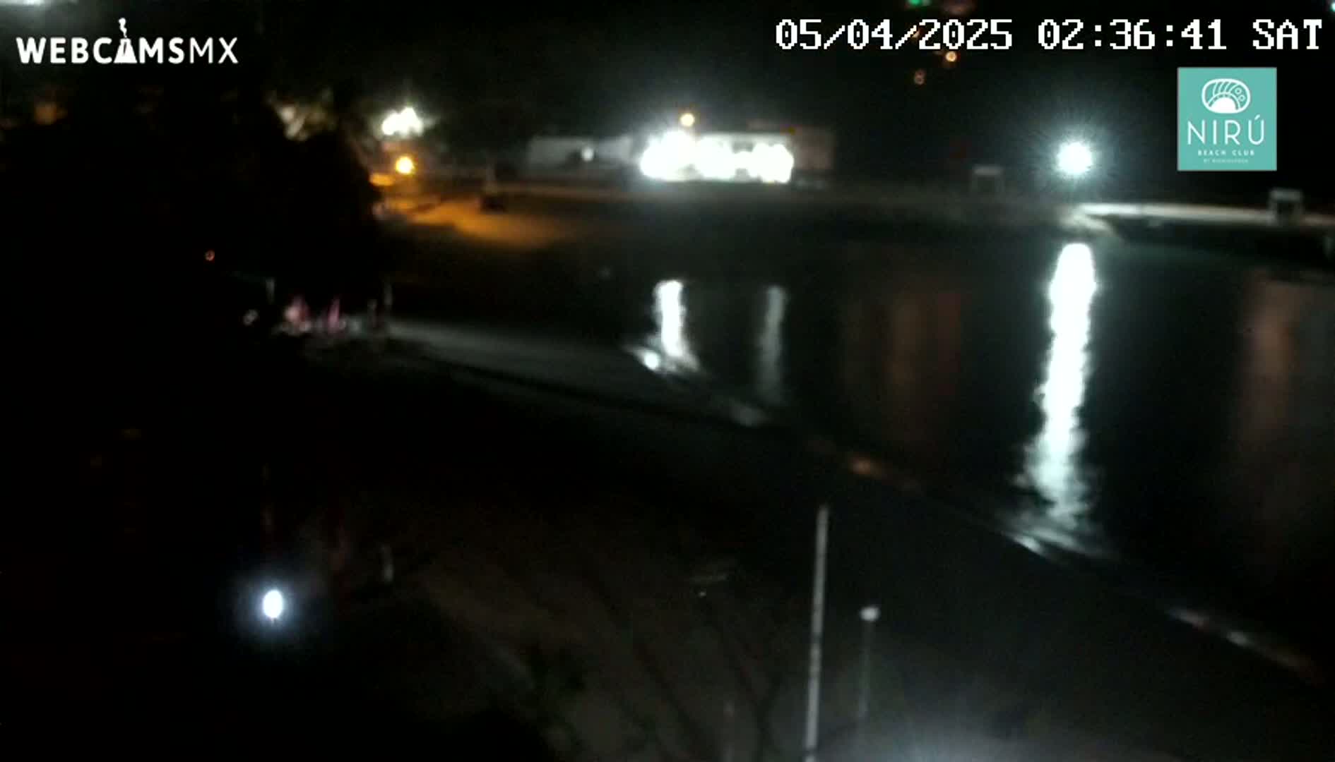 A nighttime view of a dark beach with calm water reflecting lights from a nearby roadway.