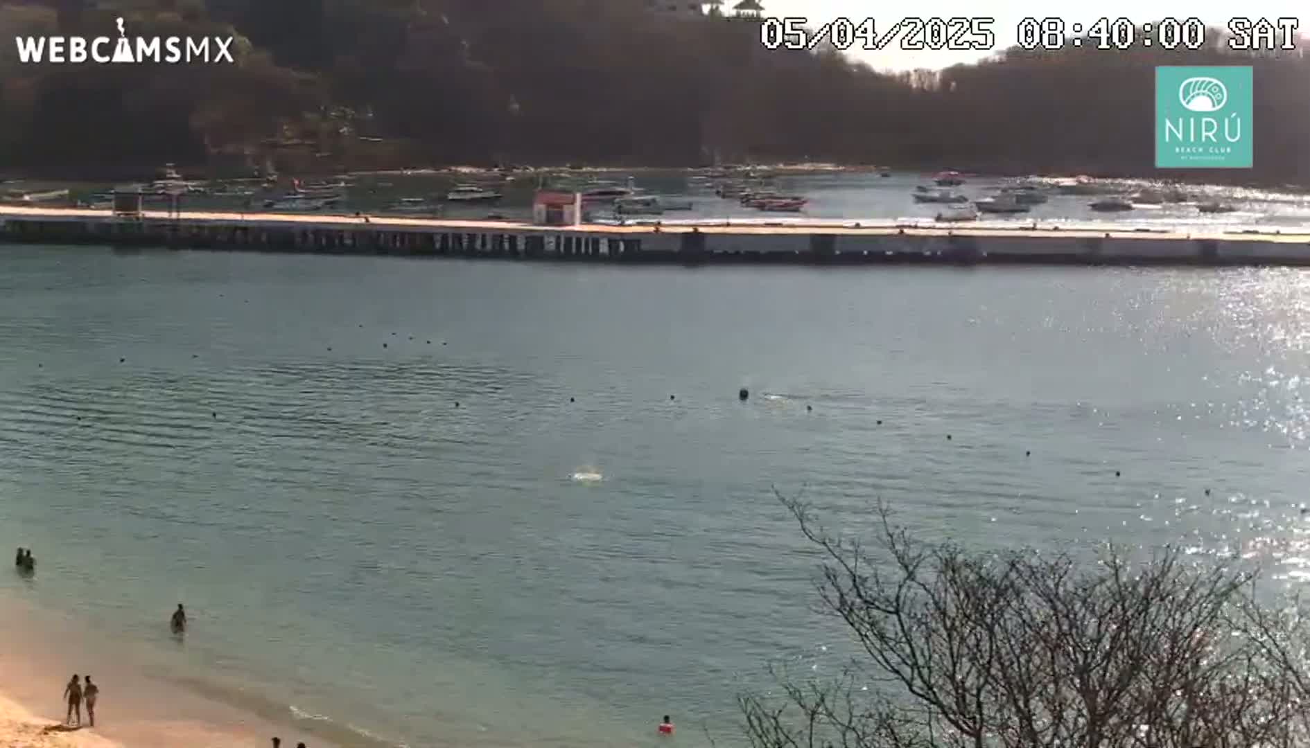 A calm, sunny day shows a long pier with boats moored alongside it, a sandy beach with a few people, and calm water extending into the distance.