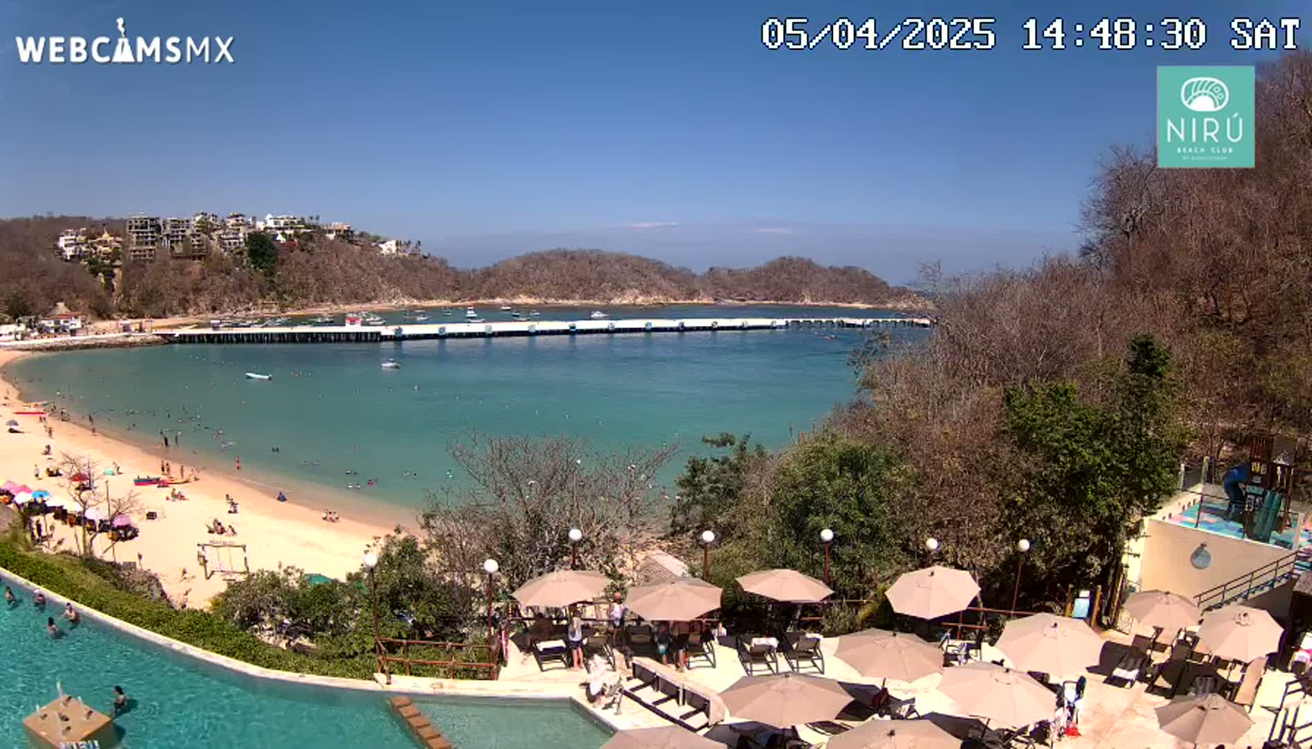 A sunny day overlooking a calm bay with a sandy beach, a long pier, and a resort pool area with many umbrellas.