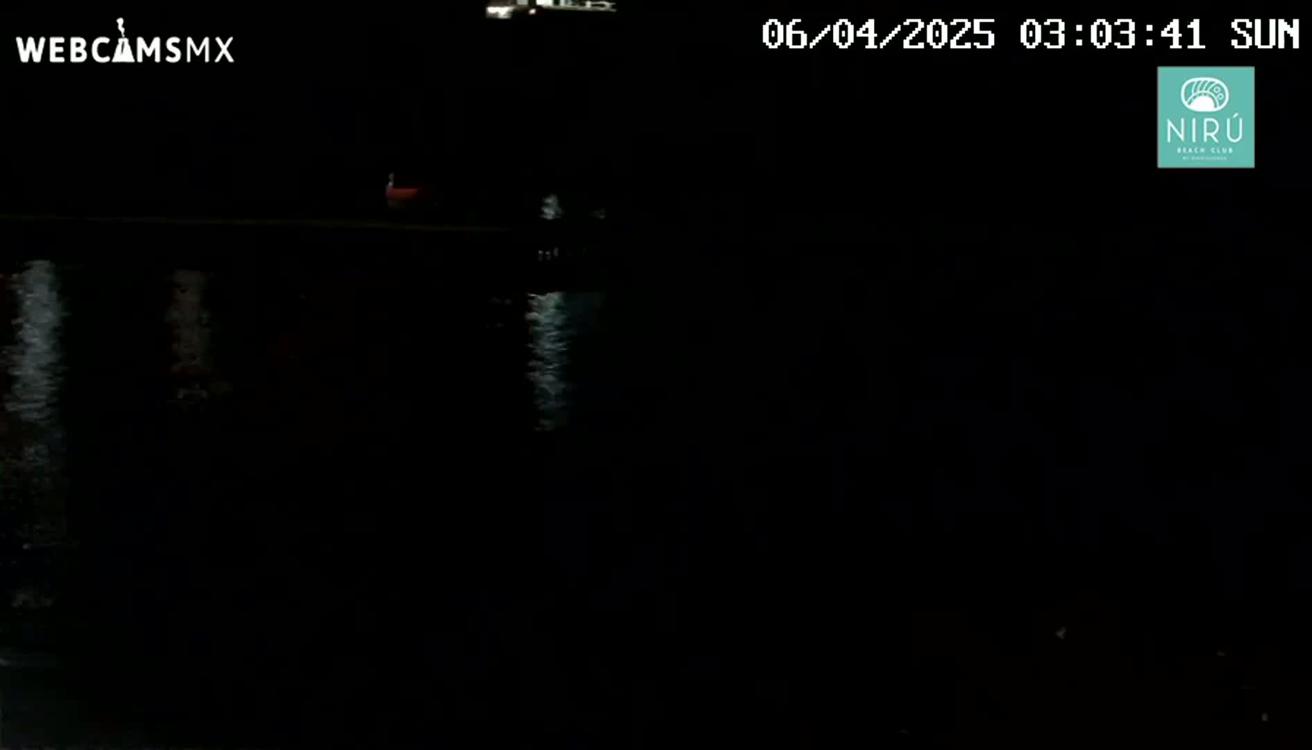 A dark nighttime scene shows a body of water reflecting faint lights, with a barely visible object near the water's edge.