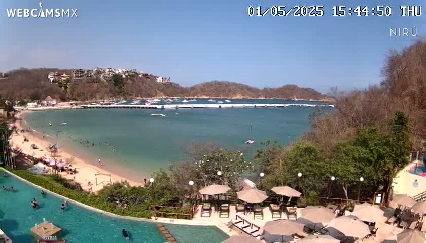 A sunny, tropical beach scene with calm, clear water, a sandy shore populated with people, and a resort area with a pool and lounge chairs under umbrellas overlooking the bay.