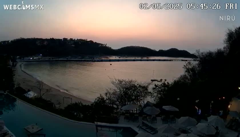 A calm bay with a sandy beach is visible at sunset, overlooking a resort area with a pool and umbrellas.