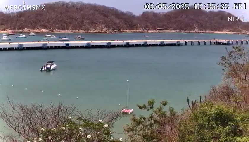 A calm, clear body of water with several boats docked at a long pier, surrounded by hillsides with sparse vegetation under a sunny sky.