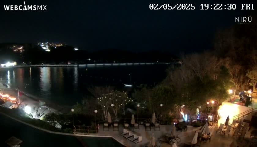 A nighttime view of a calm body of water next to a resort with outdoor seating and a swimming pool, under a clear, dark sky.