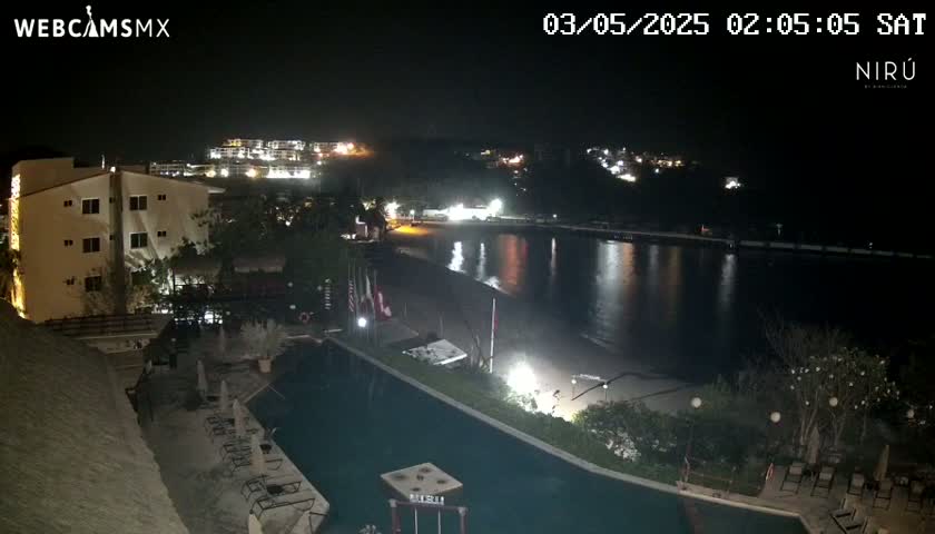 A nighttime view of a resort pool area overlooking a calm bay, with buildings and lights illuminating the landscape.