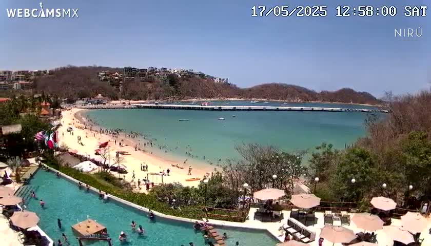A sunny day at a beachside resort shows a long pier extending into clear turquoise water, a sandy beach filled with people, and a large swimming pool overlooking the ocean.
