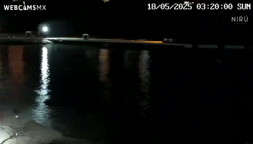 A nighttime view of a calm body of water reflecting lights from a dock under a clear, dark sky.