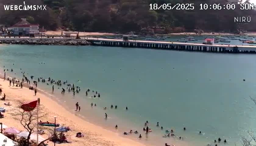 A sunny day at a beach with calm, light-blue water shows many people swimming and relaxing on the sand near a pier and a small building.