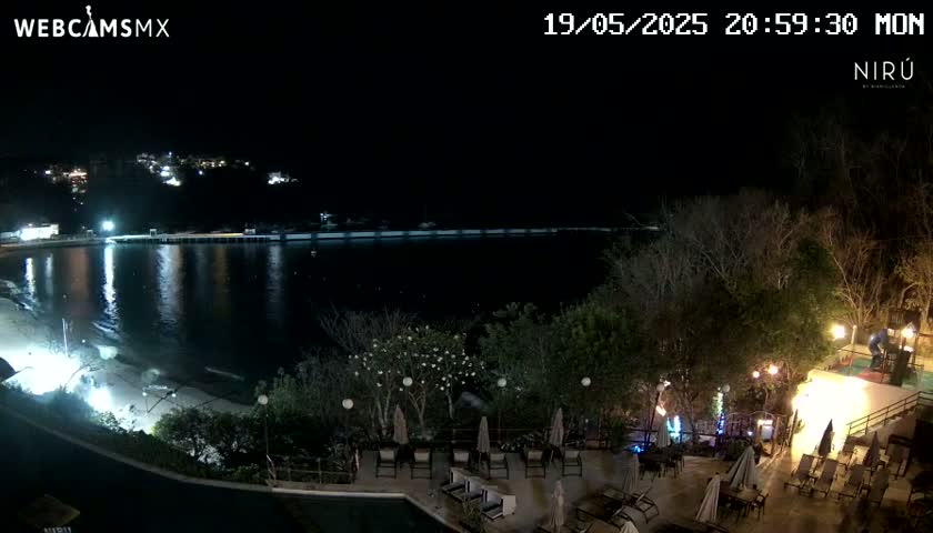 A nighttime view of a calm bay with a beach and a resort area with outdoor furniture and lighting.