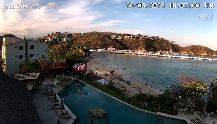 An aerial view of a resort pool and beach area, overlooking a calm bay with buildings and people on the shore under a partly cloudy sky.