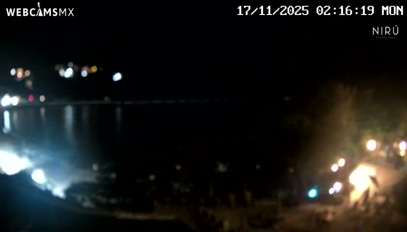 A blurry night-time view reveals numerous scattered lights reflecting on a calm body of water, with additional lights outlining distant and nearby shores under clear conditions.