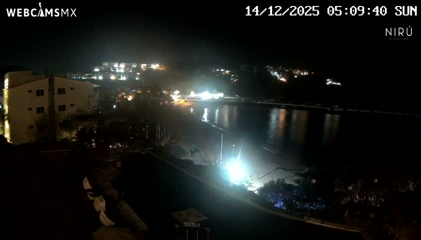 The image shows a nighttime view of a bay or ocean reflecting distant city lights and bright shore illumination, alongside a well-lit outdoor patio with tables, chairs, and umbrellas, under clear night sky conditions.