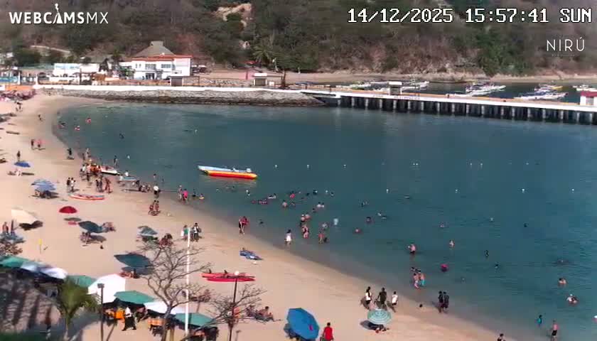 The image shows a nighttime view of a bay or ocean reflecting distant city lights and bright shore illumination, alongside a well-lit outdoor patio with tables, chairs, and umbrellas, under clear night sky conditions.