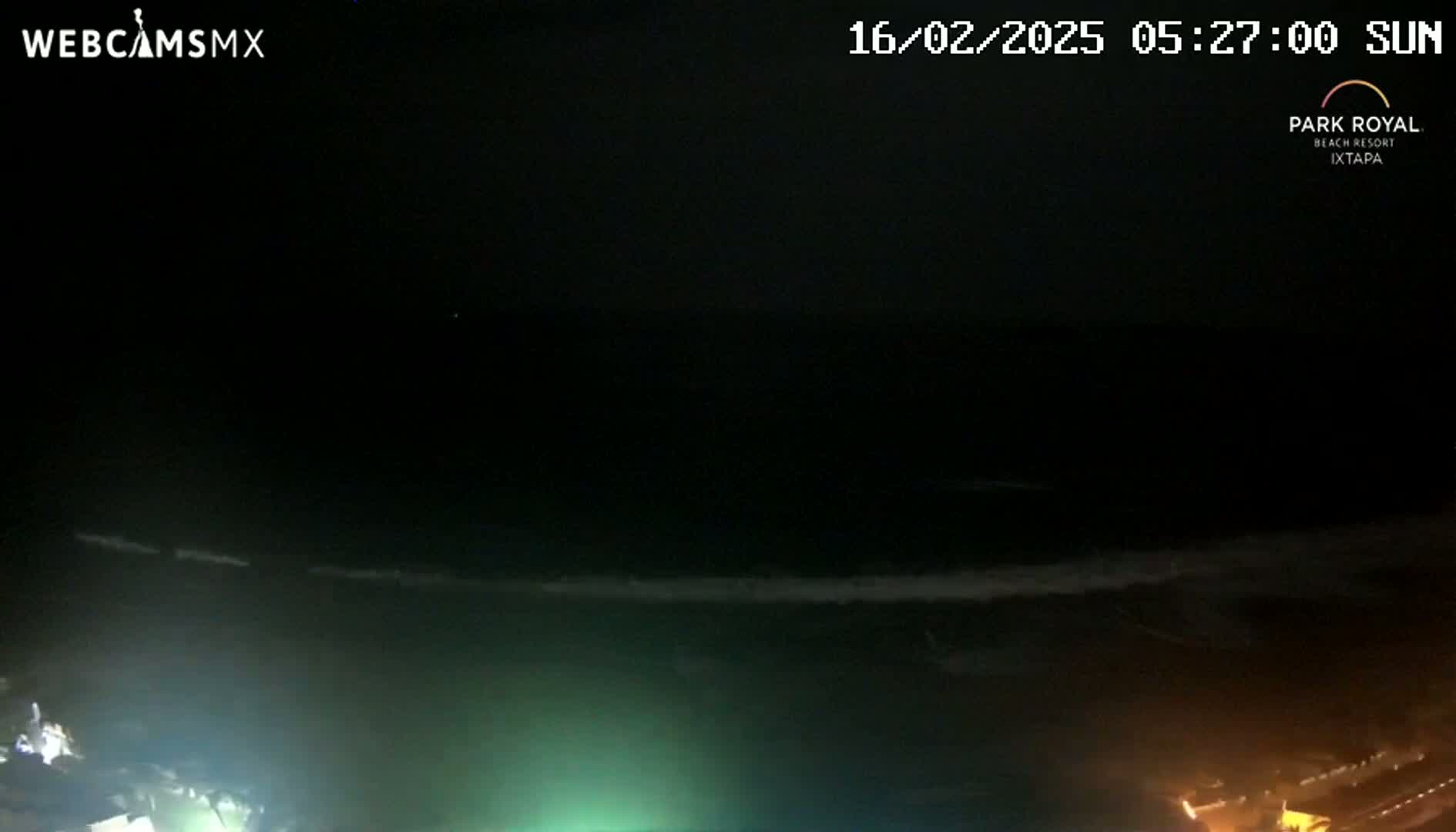 A nighttime, ocean beach scene shows dark water with small waves gently breaking near the shore, illuminated by lights on the land.