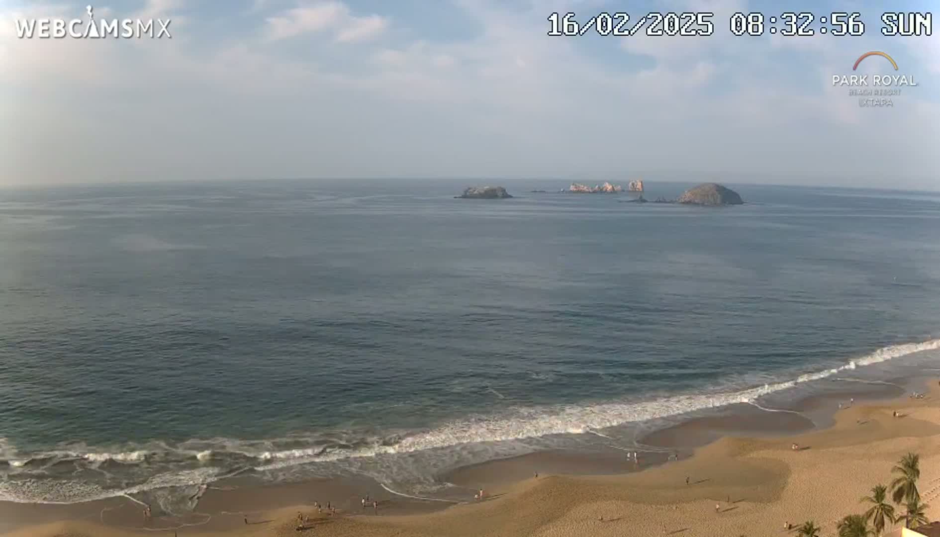A sandy beach with gentle waves laps the shore under a mostly clear sky, with several small islands visible in the distance.