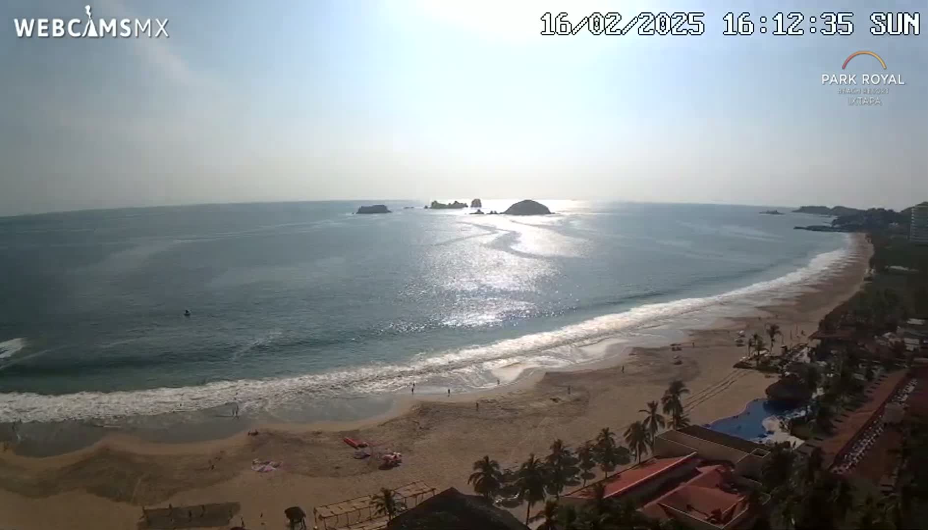 A sunny day at a beach with calm waves, showing a coastline with several buildings and palm trees, and a group of small islands in the distance.