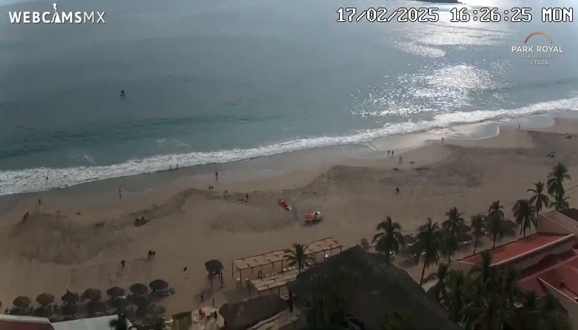 An aerial view shows a beach with calm waves, a few people scattered on the sand, and palm trees lining the beachfront buildings.