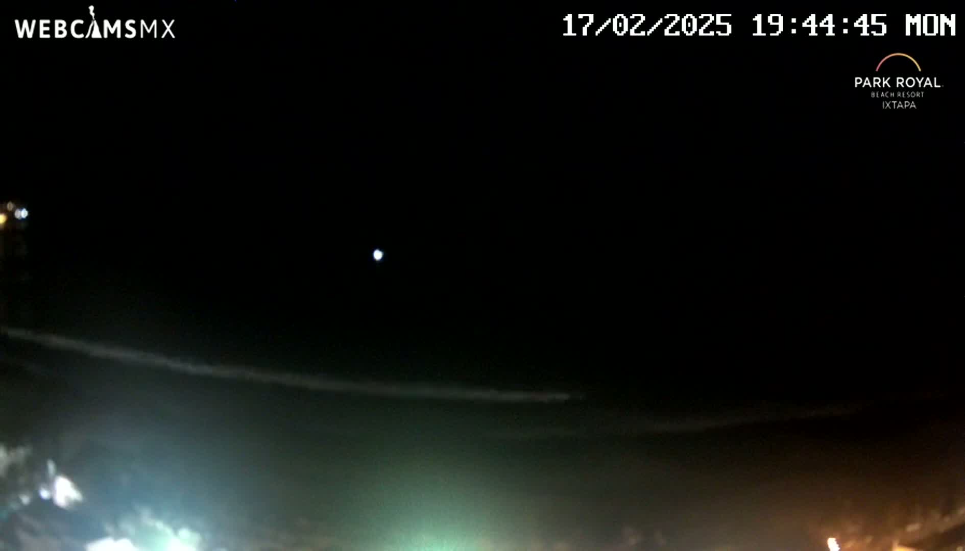 A nighttime view of a dark ocean with a faint, bright light in the sky and a shoreline with dim lights.