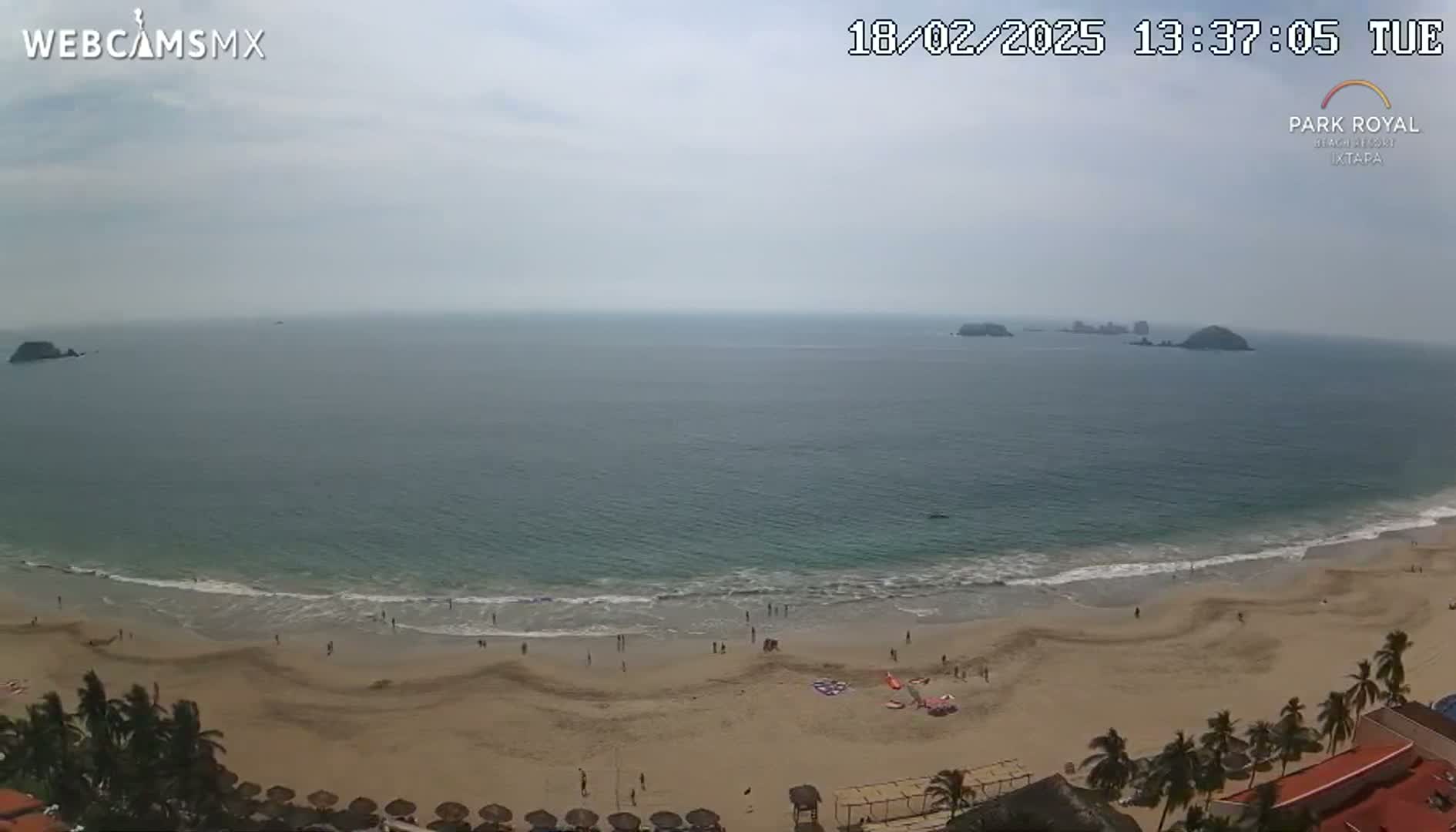 An overhead view shows a sandy beach with a few people on it, gentle waves lapping the shore, and a calm ocean extending to distant islands under a mostly cloudy sky.