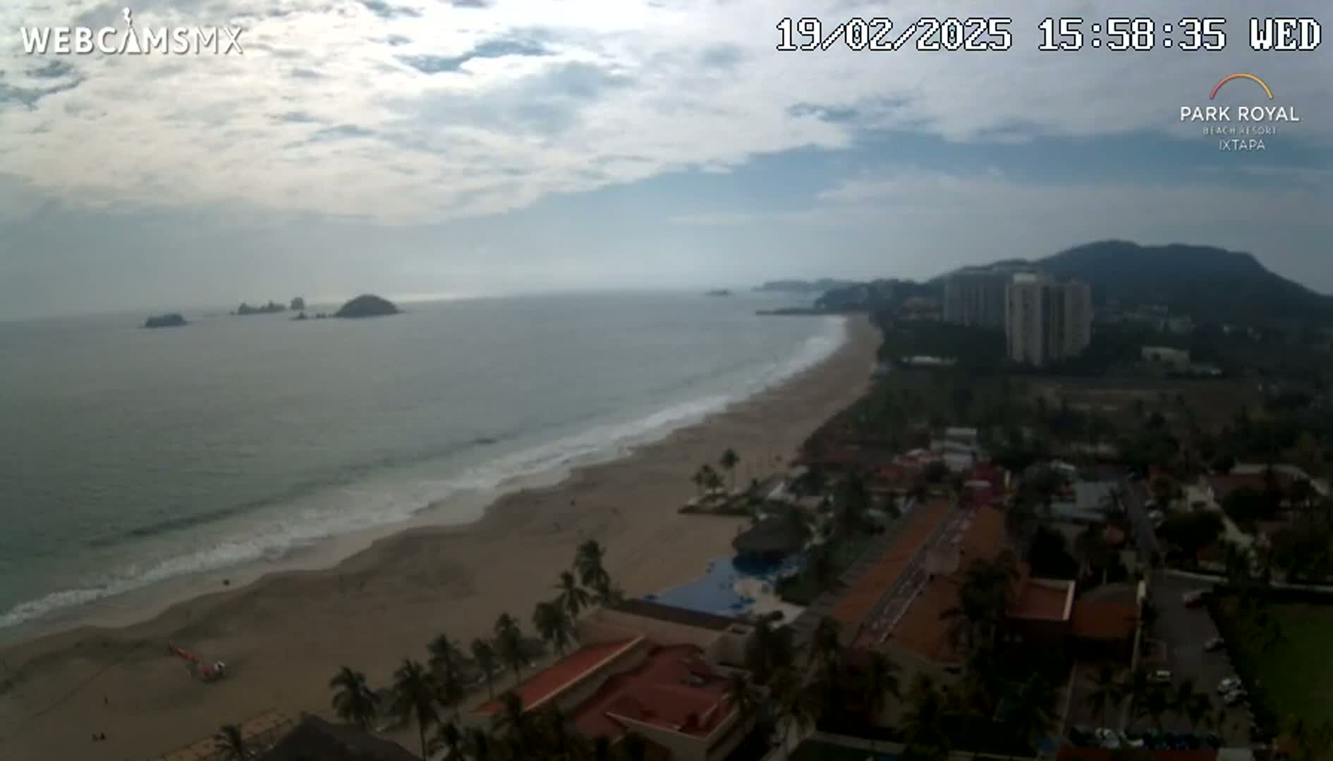 An ocean beach with waves lapping a sandy shore, backed by hotels and high-rise buildings under a mostly cloudy sky.