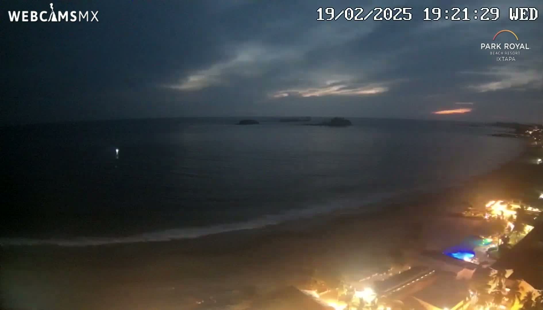 A nighttime coastal scene shows a dark ocean with small waves lapping a beach, some distant islands, and a stretch of illuminated beachfront property under a mostly cloudy sky.