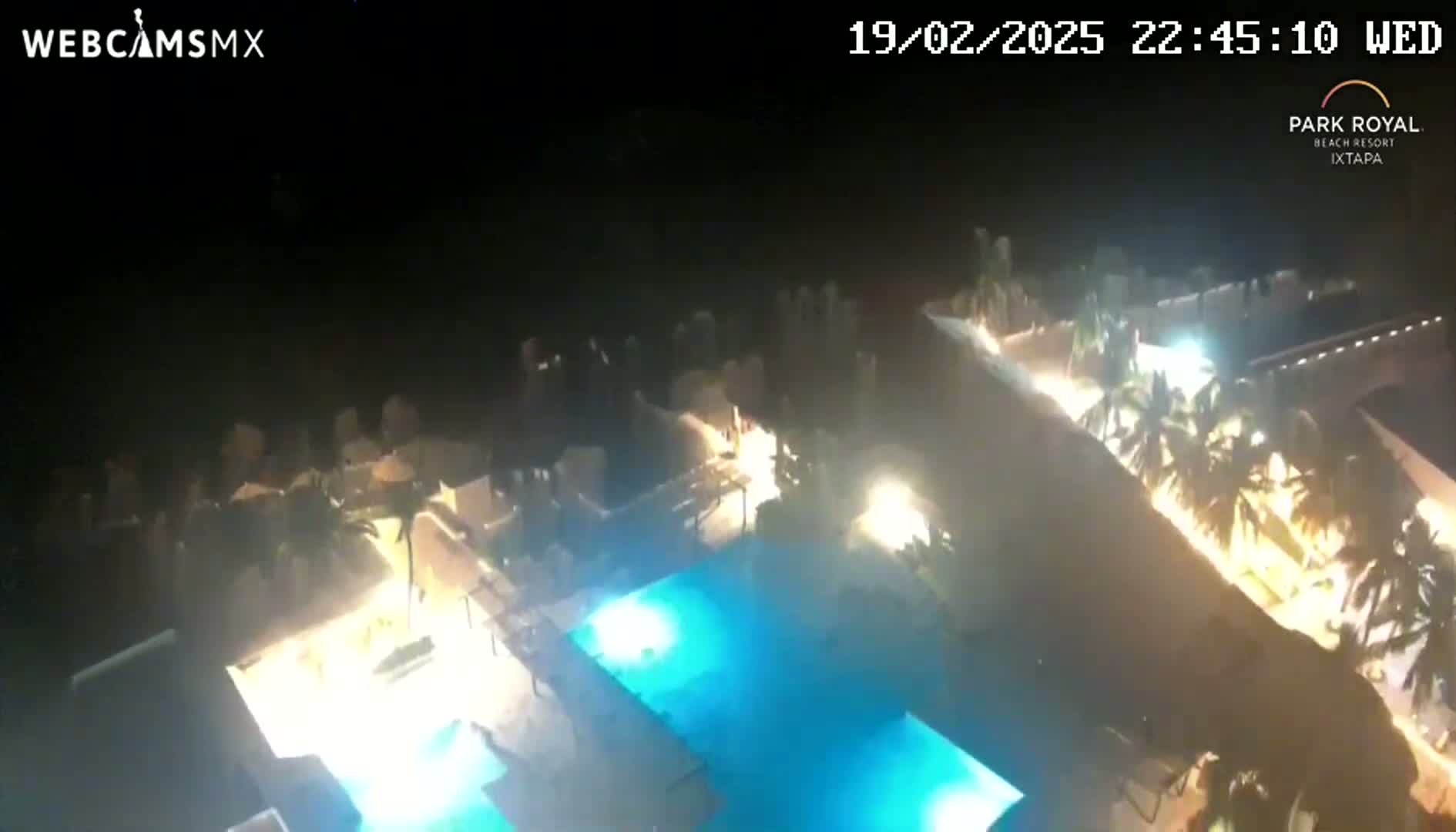 An aerial nighttime view shows a resort with illuminated swimming pools and palm trees under a clear, dark sky.
