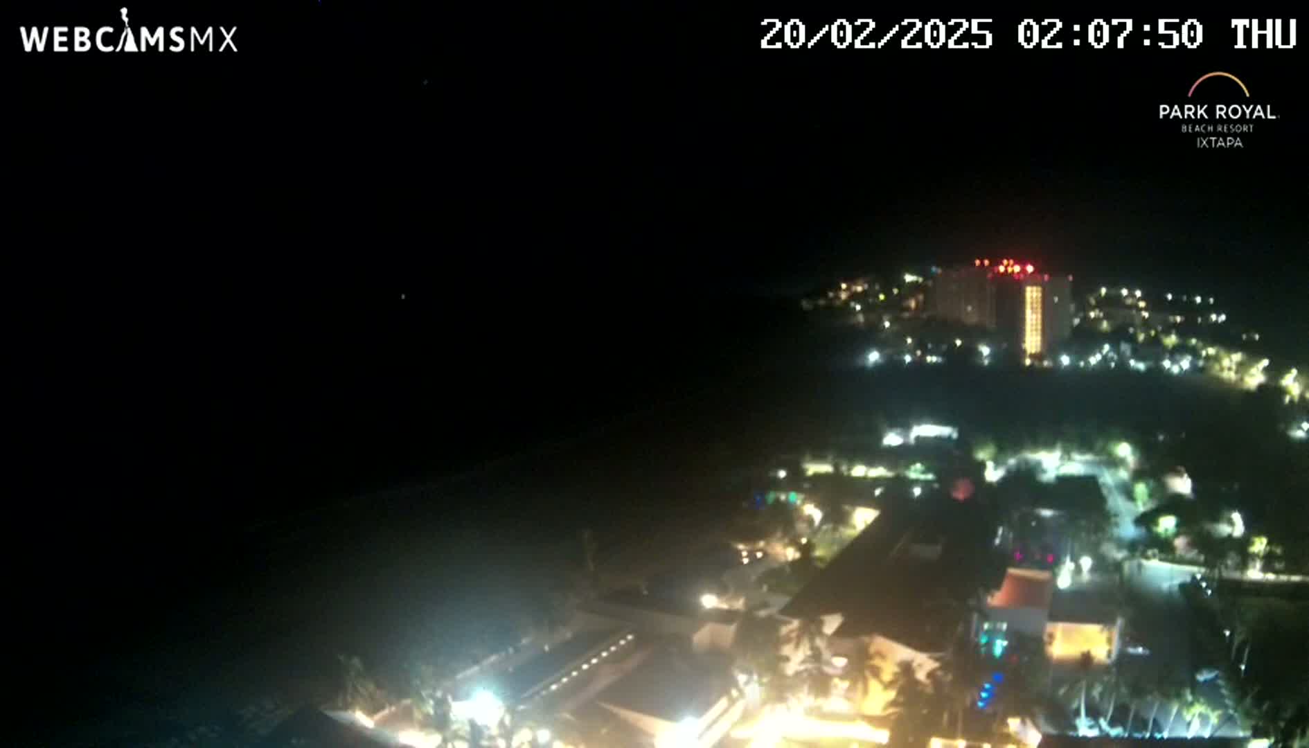A nighttime, high-angle view shows a resort town with numerous lights illuminating buildings and landscaping next to a dark body of water under a clear, dark sky.