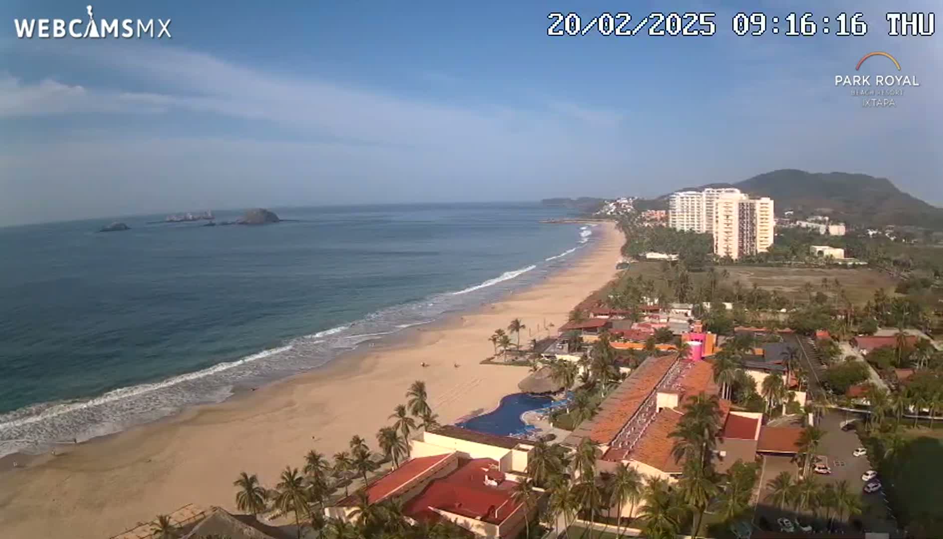 An aerial view shows a sandy beach with gentle waves lapping the shore, lined with palm trees and resort buildings under a mostly sunny sky.