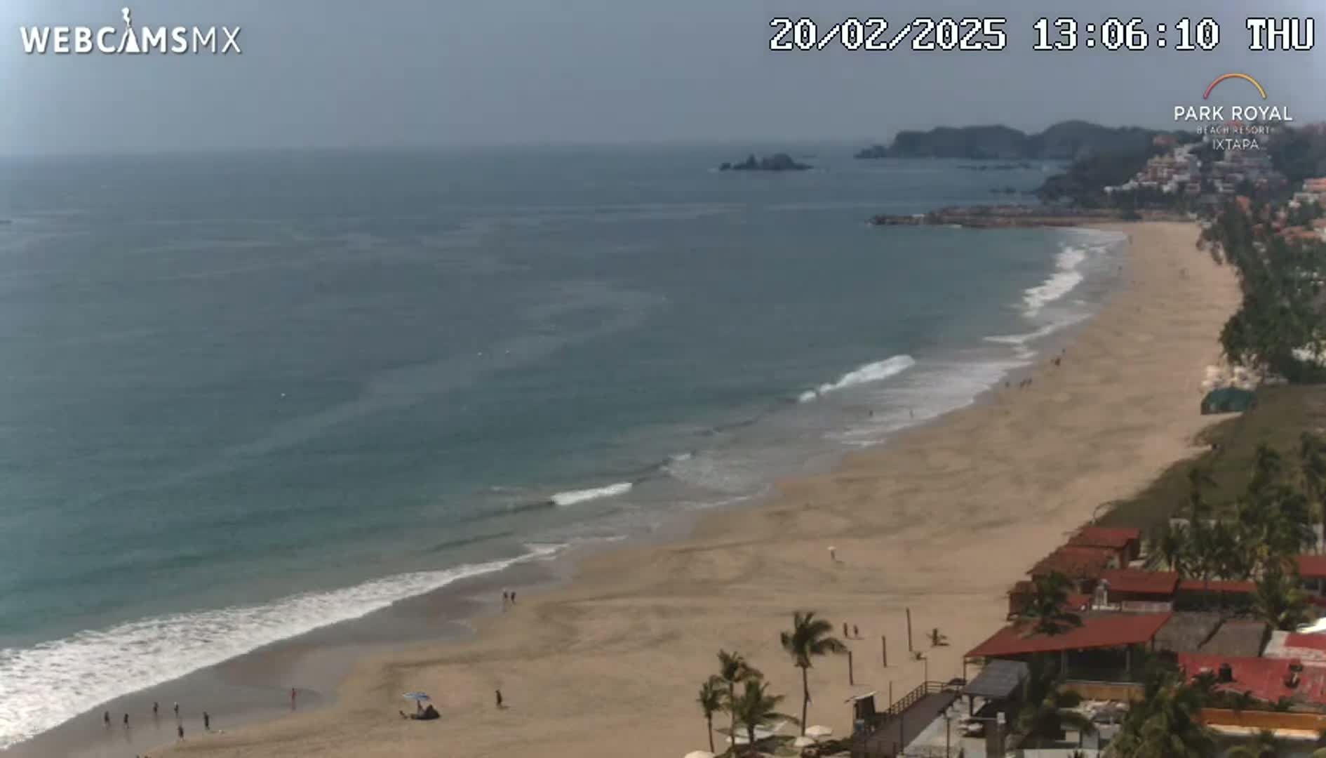 A long sandy beach with gentle waves and a few people on it, next to buildings and palm trees under a mostly clear sky.