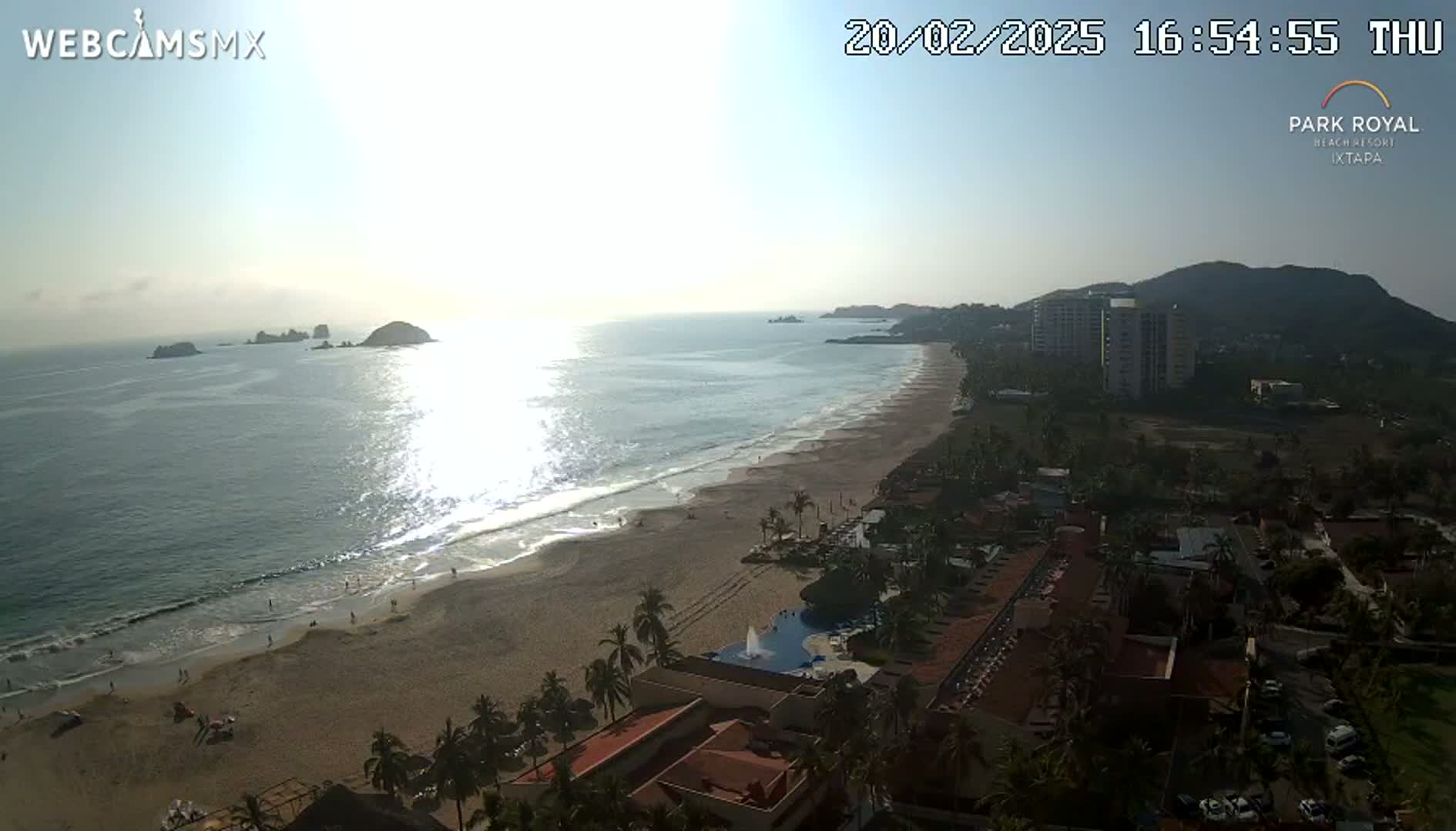 An aerial view shows a beach with calm waves under a sunny sky, palm trees, and resort buildings along the coastline with a mountain in the background.