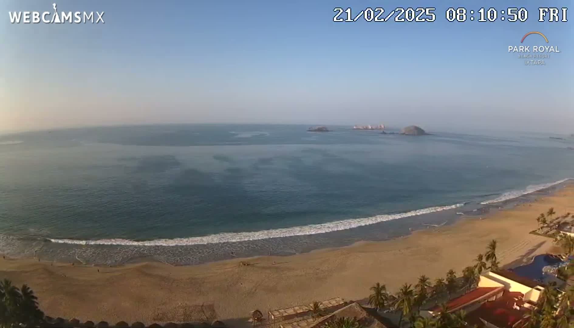 A sandy beach with gentle waves meets a calm ocean under a clear, sunny sky, with islands visible in the distance.