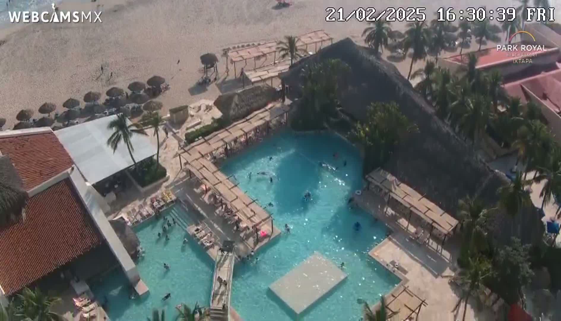 An aerial view shows a resort pool complex with many people swimming and lounging, adjacent to a sandy beach on a sunny day.