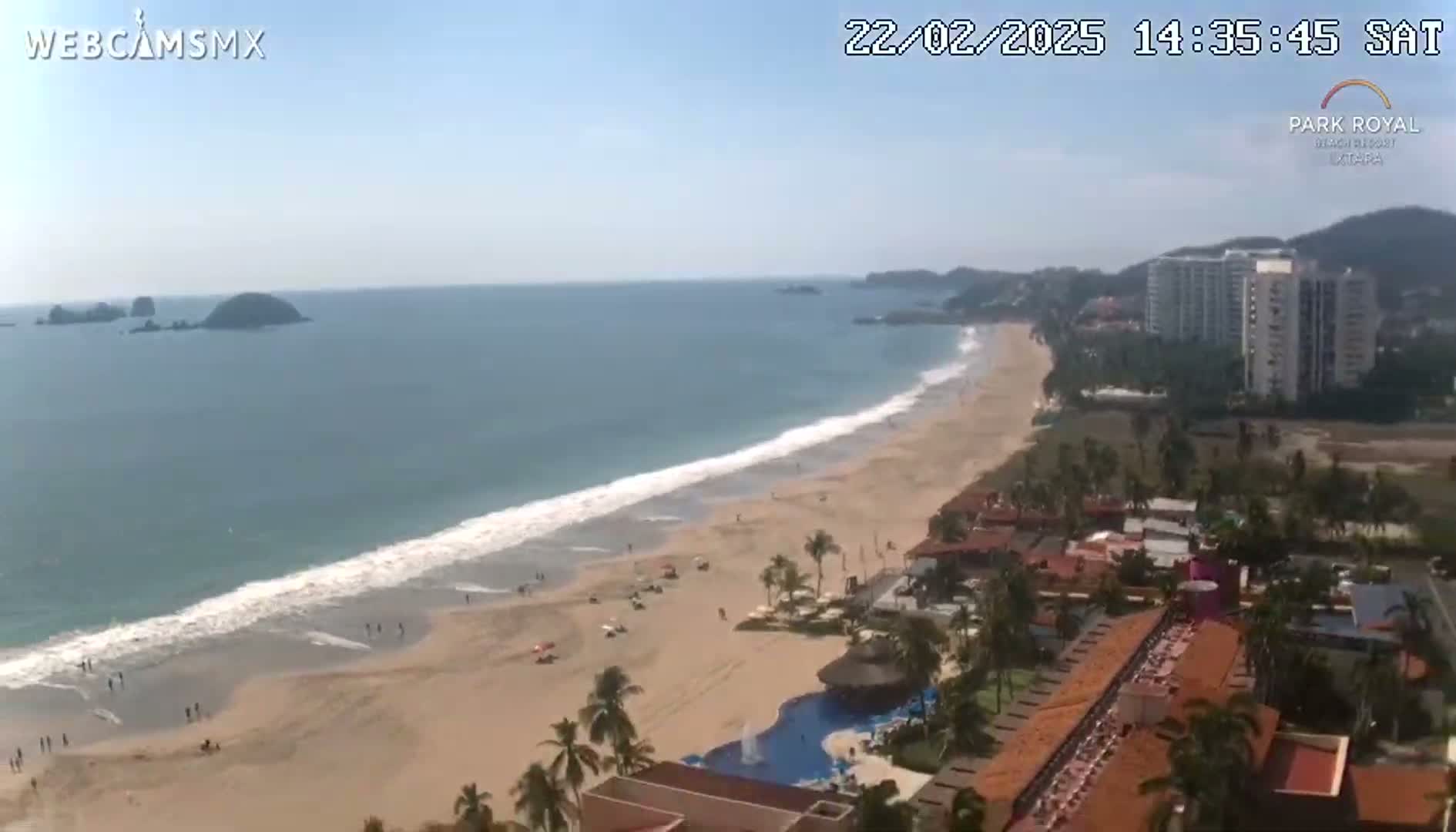 A sandy beach with gentle waves laps the shore, overlooking a resort with a pool and high-rise buildings under a clear, sunny sky.