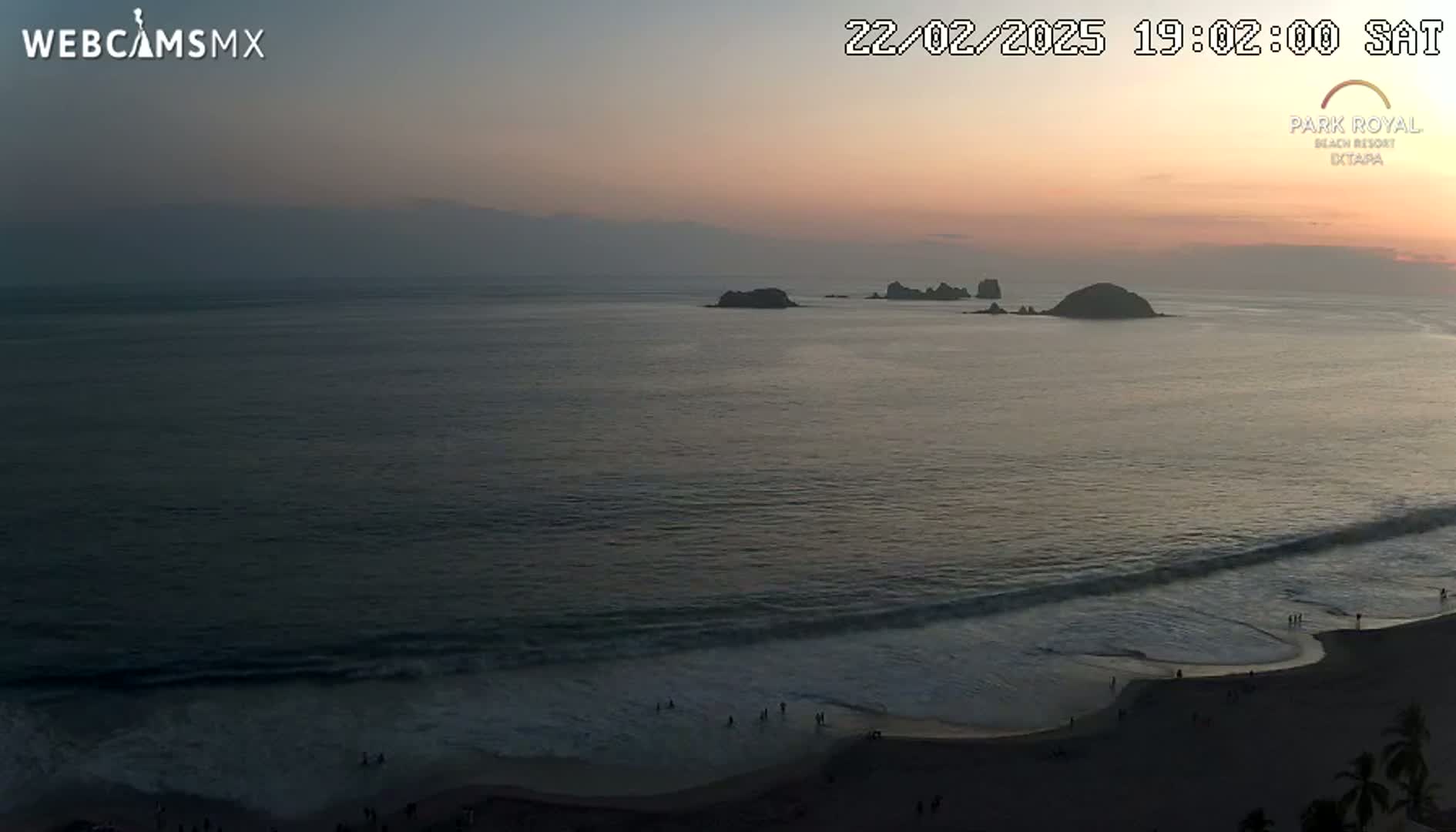 A tranquil ocean scene at sunset shows a beach with a few people, gentle waves rolling onto the shore, and several small islands in the distance.