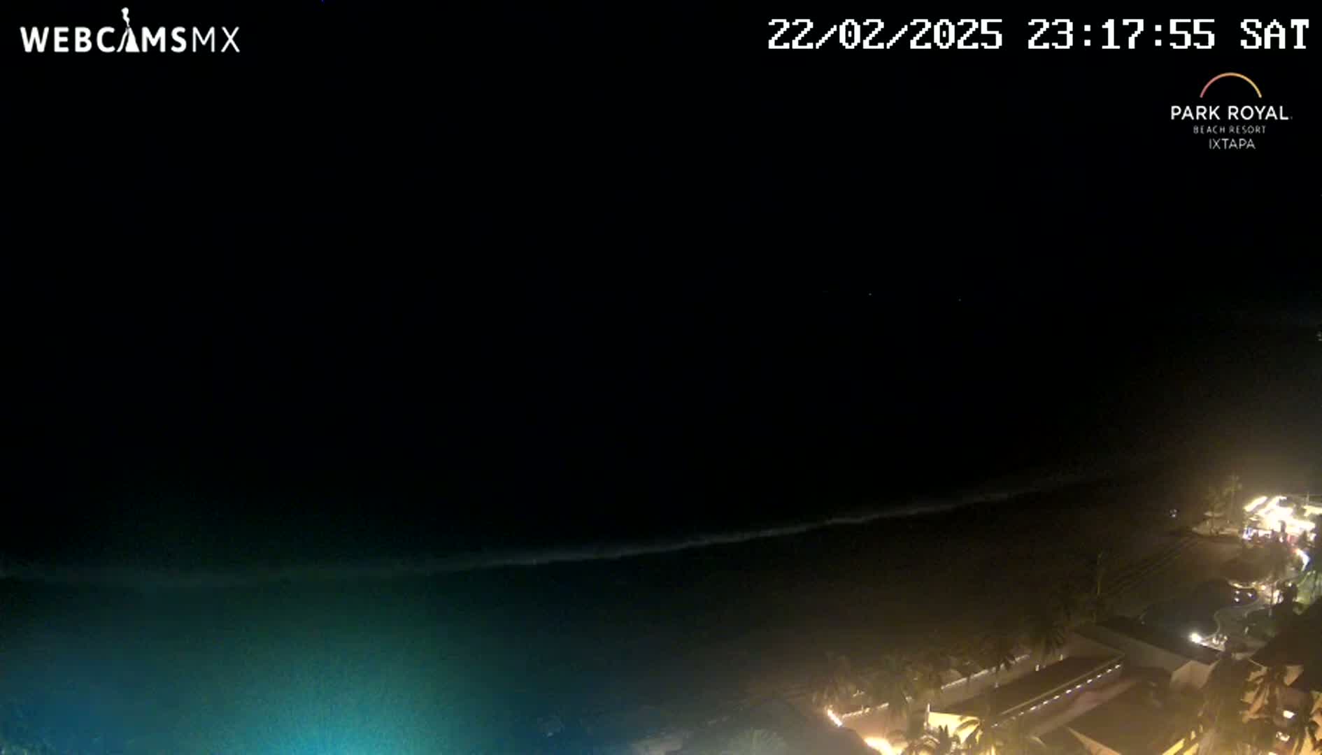 A nighttime coastal scene shows dark water with a faint line of waves, and illuminated buildings and vegetation along the shore under a clear, dark sky.