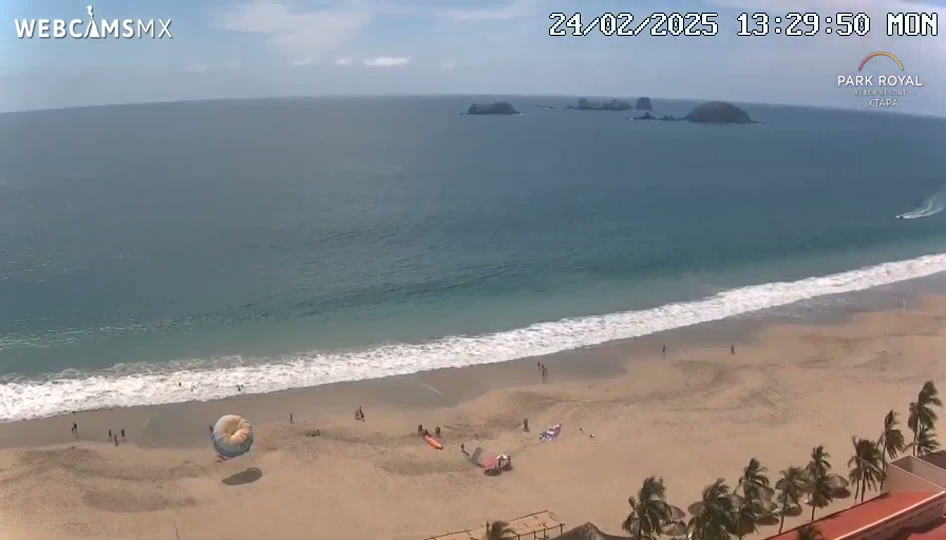 A sandy beach with a few people and a parasail is visible, with calm ocean waves lapping the shore under a partly sunny sky.