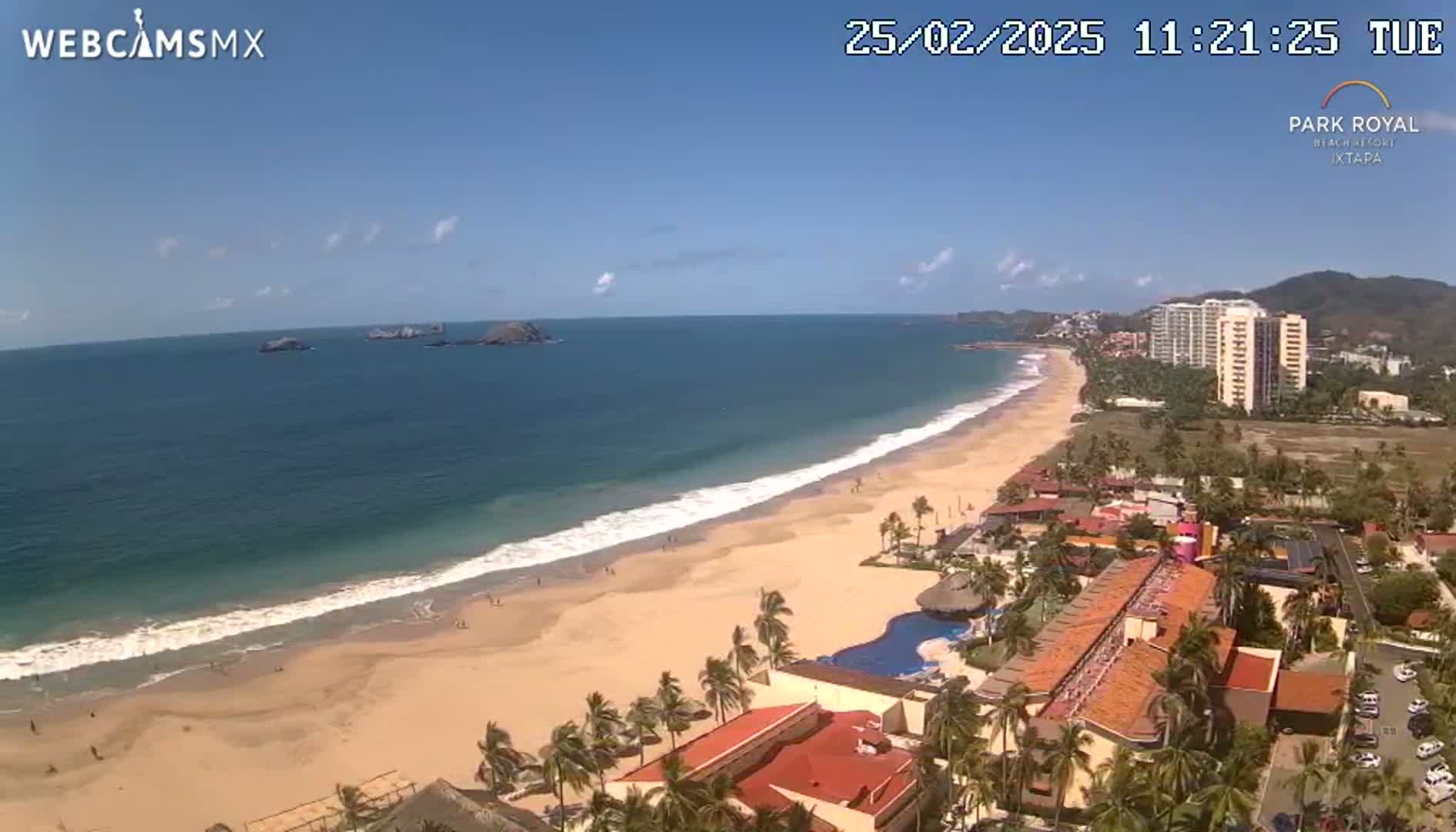 An aerial view shows a long sandy beach with gentle waves lapping the shore under a clear blue sky, featuring beachfront hotels and palm trees.