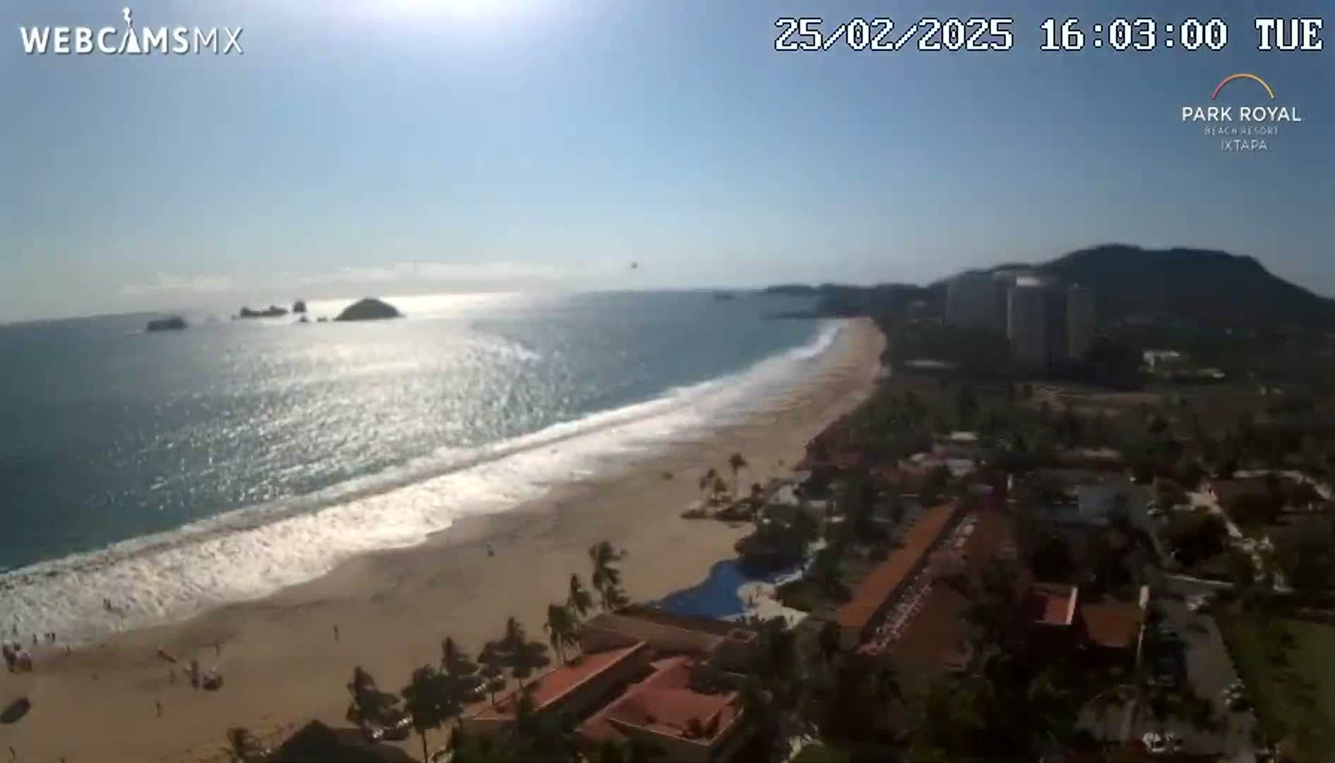 An aerial view shows a sunny beach with gentle waves, a resort complex, and distant mountains under a clear sky.