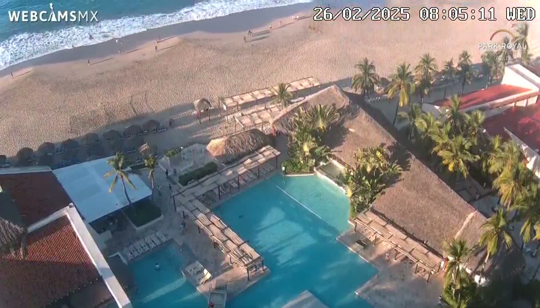 An aerial view of a resort pool area adjacent to a sandy beach with light ocean waves under sunny skies.