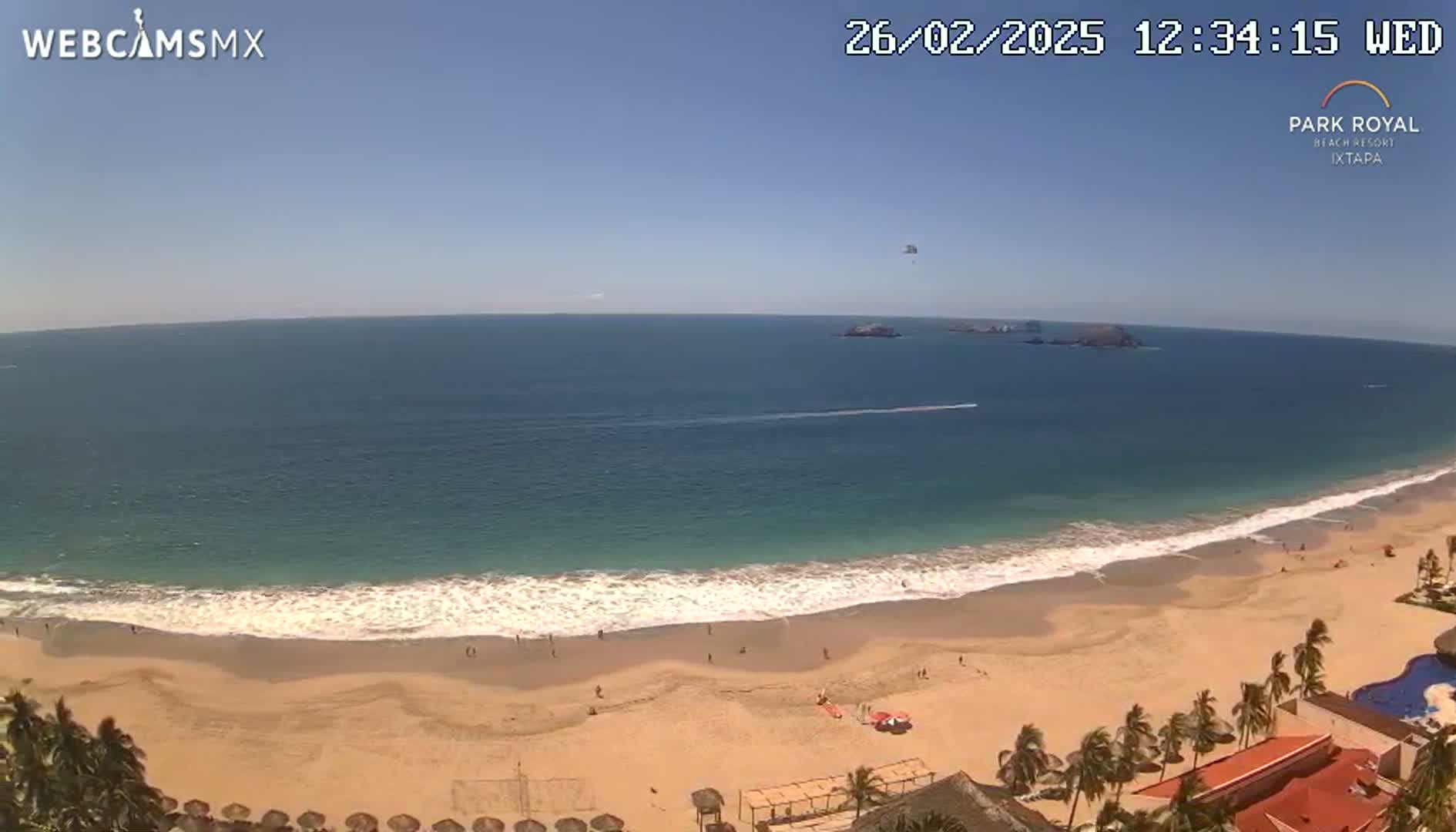 A beach with calm waves under a clear blue sky, a few people are visible on the sand, and parasailors are in the distance over the ocean.