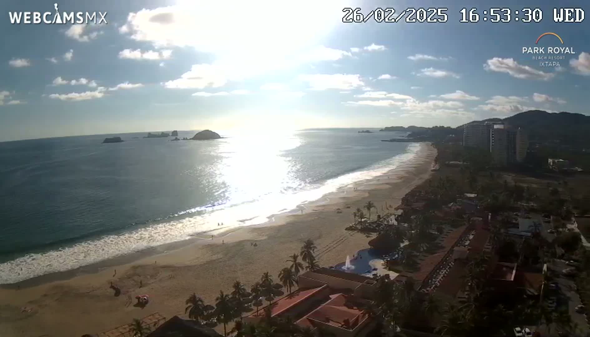 A sunny day shows a long sandy beach with gentle waves lapping the shore, overlooking a resort and ocean with islands in the distance.