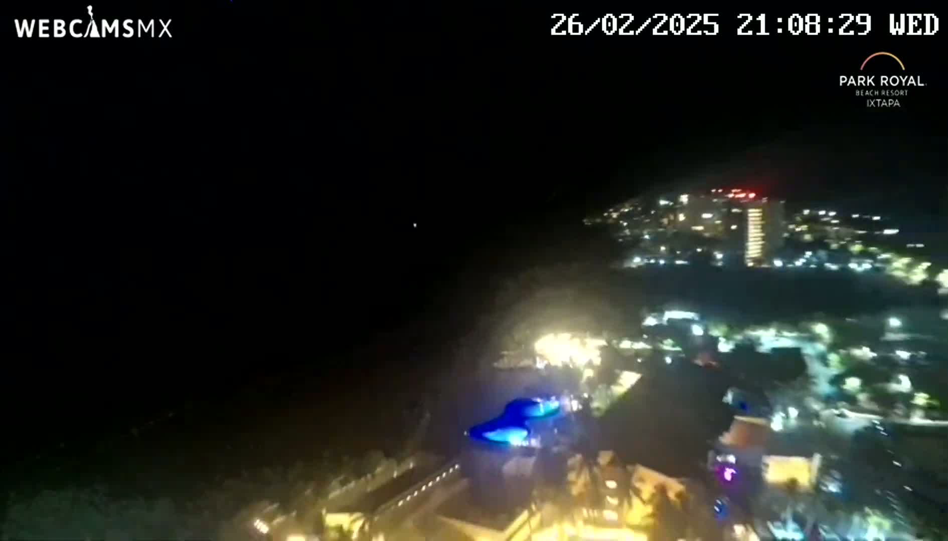 A nighttime aerial view shows a brightly lit resort area next to a dark ocean under a clear sky.