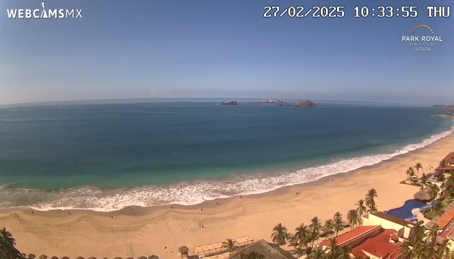 A sandy beach with gentle waves and a clear blue sky stretches out to the ocean, with palm trees and buildings visible along the shore.
